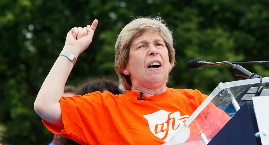 Randi Weingarten in an orange shirt with the AFT logo gestures angrily behind a podium