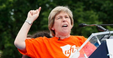 Randi Weingarten in an orange shirt with the AFT logo gestures angrily behind a podium