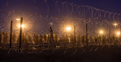 Migrants line up behind a border fence at night under bright lights