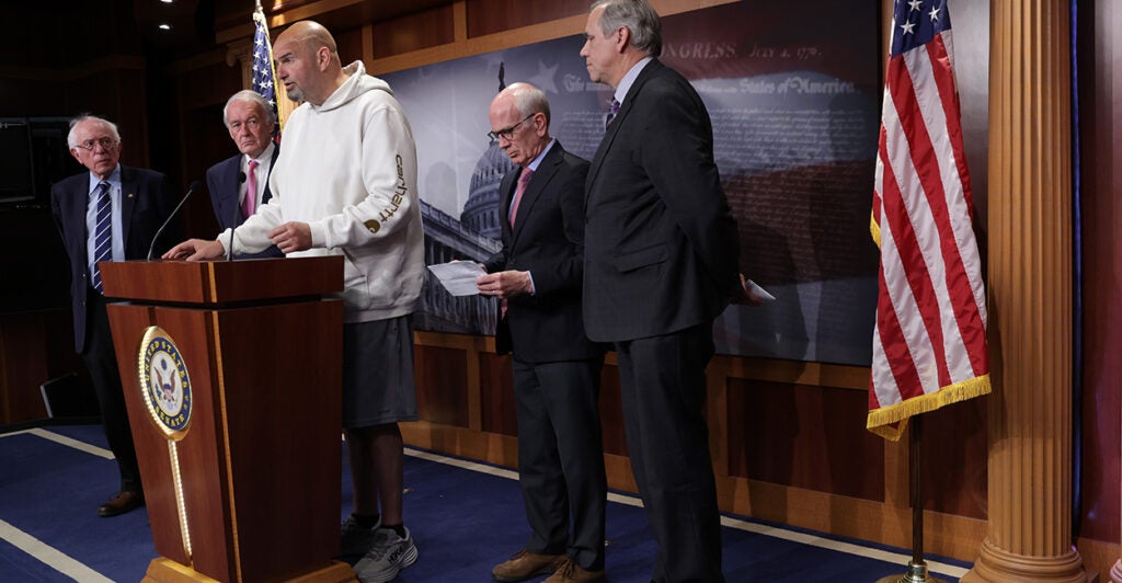 John Fetterman stands behind a congressional podium wearing a white hoodie, grey shorts and grey running shoes. Bernie Sanders, Ed Markey, Peter Welch, and Jeff. Merkley stand behind him.