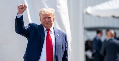 President Donald Trump greets a crowd raising his arm, wearing a black suit and a read tie