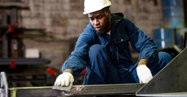 Metal worker in blue coverall and hardhat sands the surface of a construction frame in a metal factory.