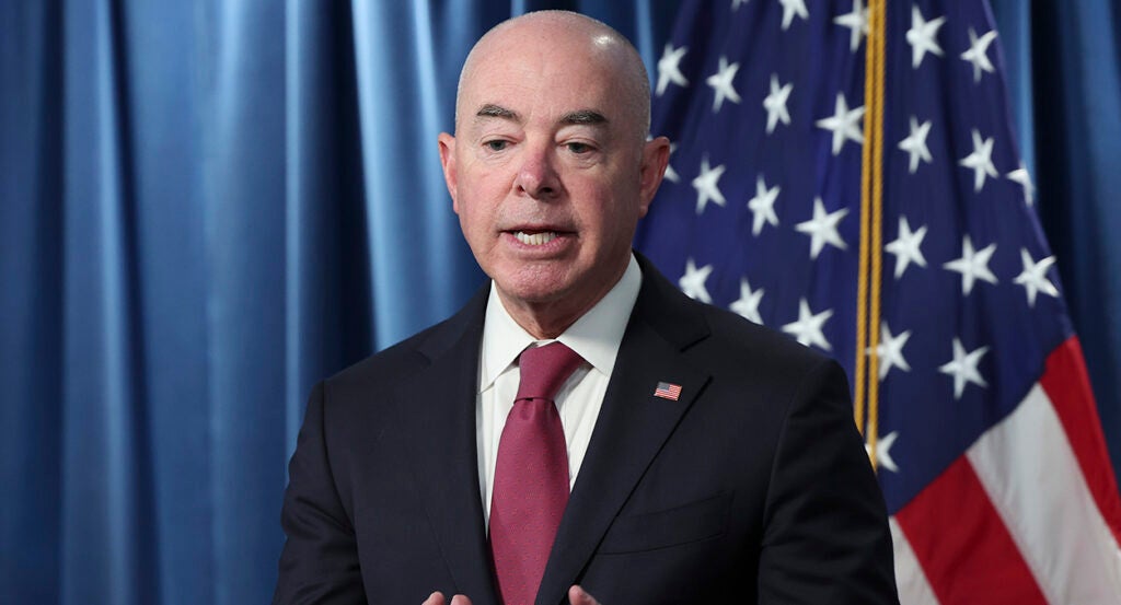 Alejandro Mayorkas in a suit with an American flag pin speaks in front of an American flag