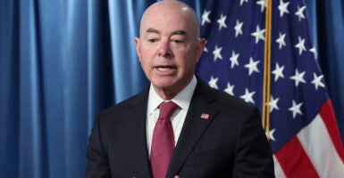 Alejandro Mayorkas in a suit with an American flag pin speaks in front of an American flag