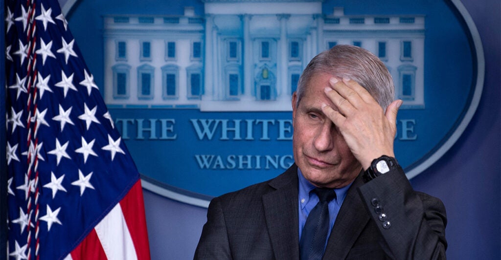 Anthony Fauci at the White House podium with his hand on his forehead