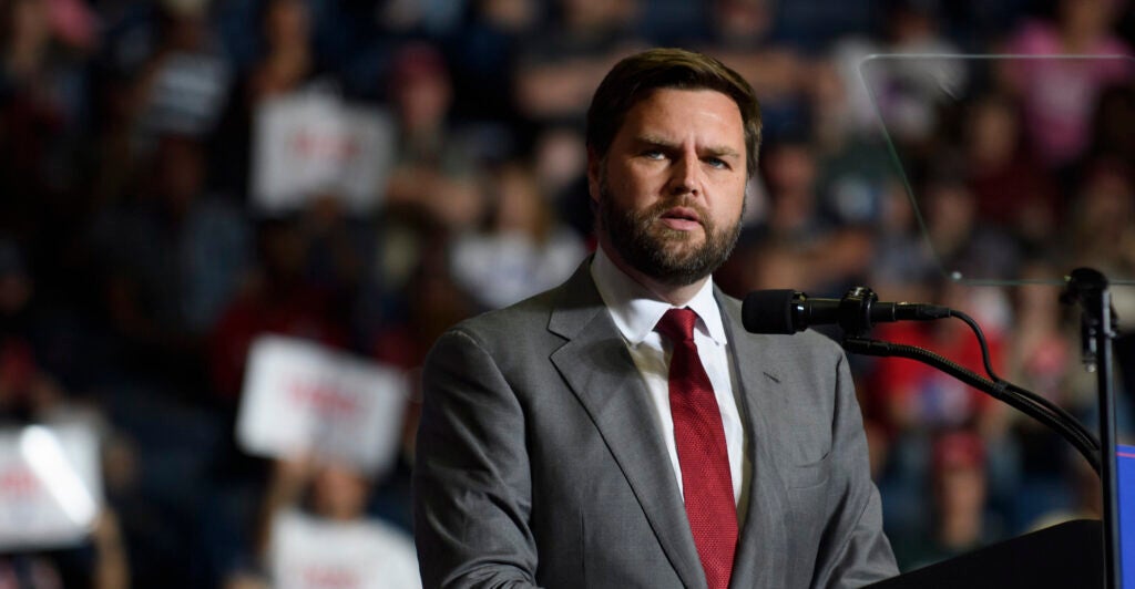 J.D. Vance in a gray suit with a red tie speaks in front of a crowd