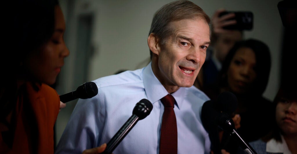 Jim Jordan in a blue dress shirt speaks in front of microphones