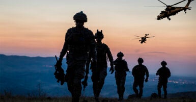 Five Army soldiers on the ground and two helicopters in the air at dusk