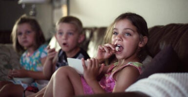 From left to right, a girl, a boy, and a girl sit on a couch eating popcorn