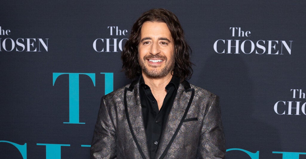 Actor Jonathan Roumie smiles on the red carpet in a grey jacket and a black shirt.