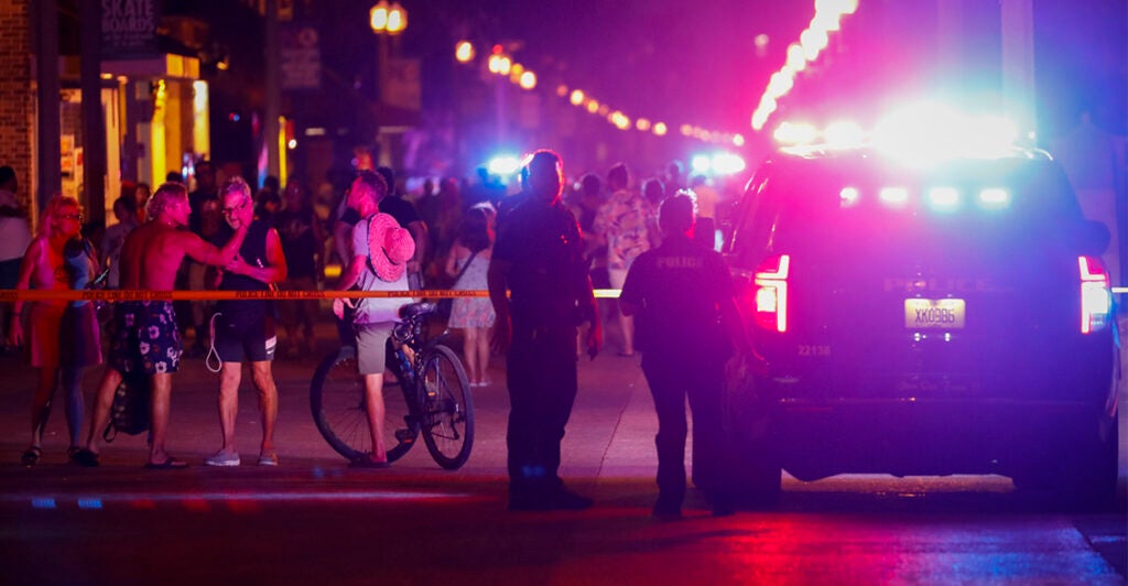 Beachgoers are seen next to law enforcement officers on a crime scene at night with police cars with lights flashing.