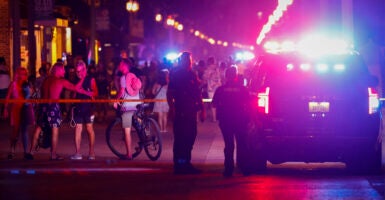 Beachgoers are seen next to law enforcement officers on a crime scene at night with police cars with lights flashing.