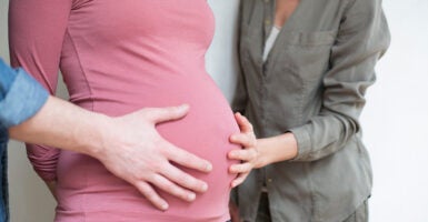 W husband and wife place a hand on the belly of the surrogate carrying their child.