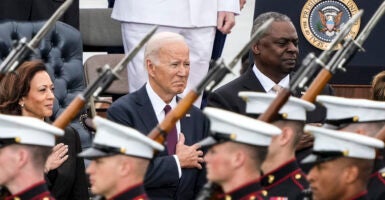 Kamala Harris, Joe Biden, and Lloyd Austin in suits stand in front of soldiers carrying rifles with bayonets.