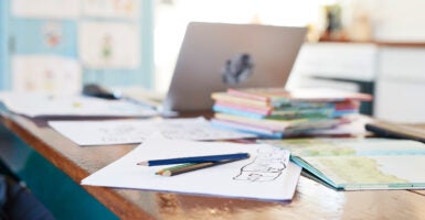 Paper, colored pencils, books, and a laptop sit on a kitchen table.