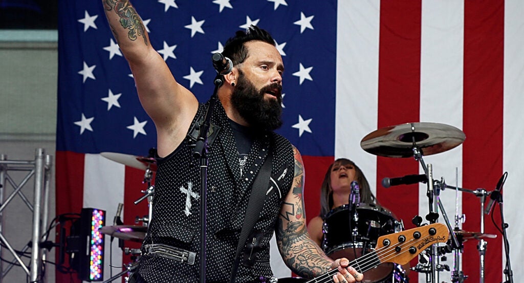 Skillet's John Cooper raises his hand while playing guitar and singing in front of an American flag.