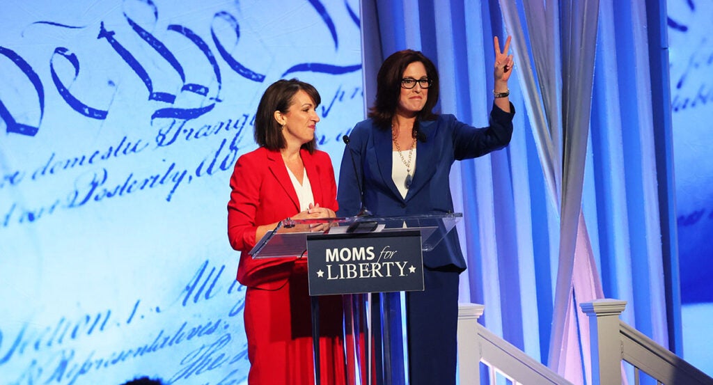 Tina Descovich in red and Tiffany Justice in blue stand before a podium with the Moms for Liberty logo