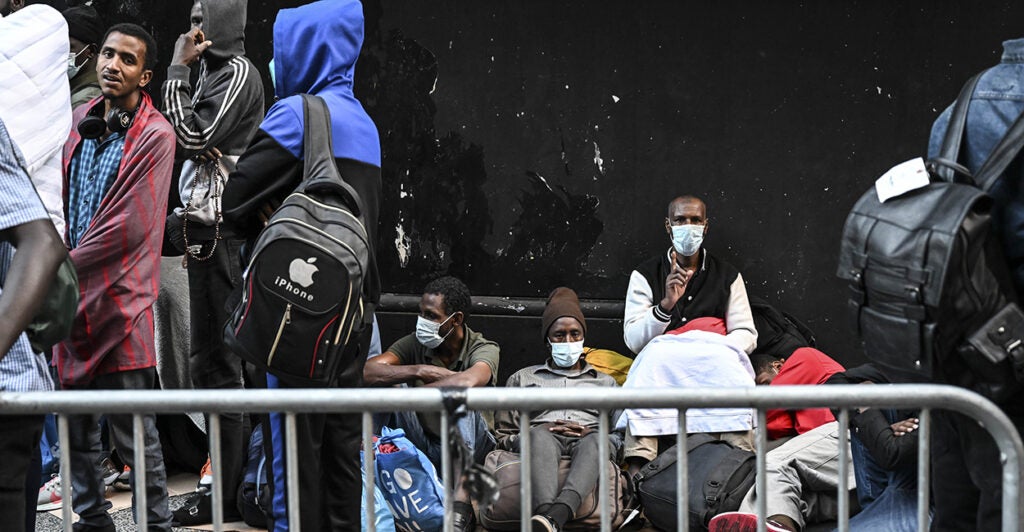 Illegal aliens sit and stand by a black wall outside a hotel in New York City.