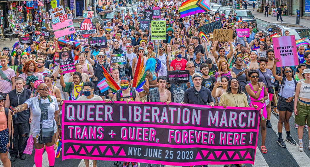 Activists in rainbow flags and various states of dress march with a "Trans and Queer" sign