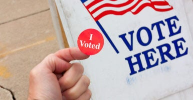 A hand holding an "I Voted" sticker with a "Vote Here" sign in the background