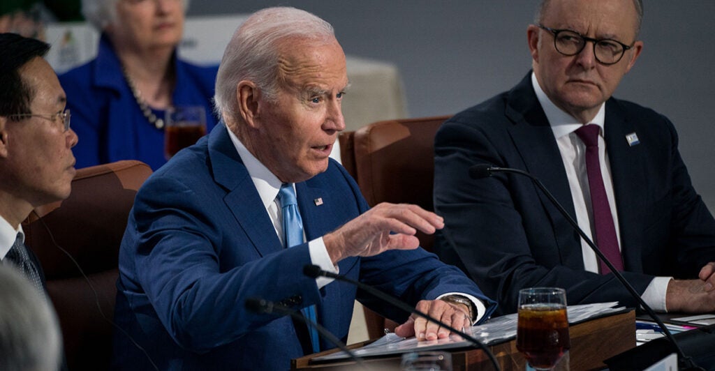 President Joe Biden sits at a table with others, speaking into a microphone