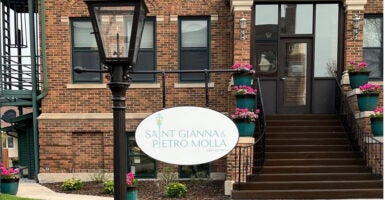 The St. Gianna and Pietro Molla Home sign hands on a lamp post outside the large brick home.