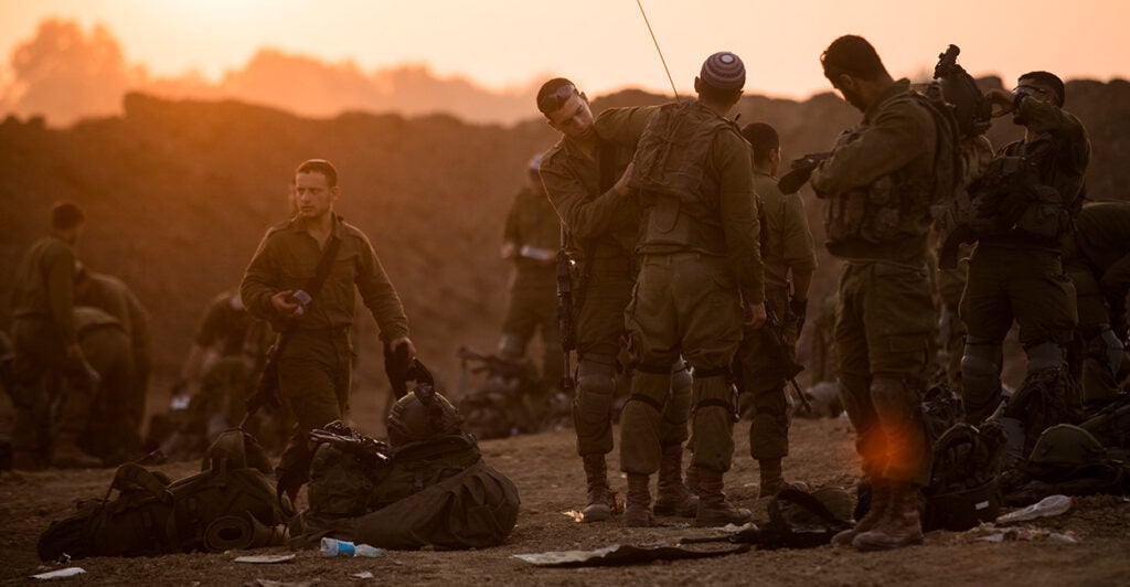 A group of Israeli troops organizes equipment on the ground as the sun peeks over a hill.