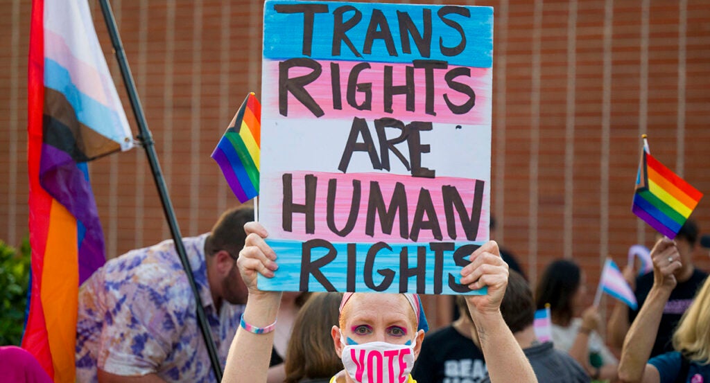 Woman holds sign reading "trans rights are human rights" while wearing a face mask reading "Vote"