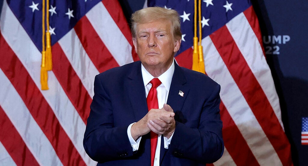 Donald Trump in a blue suit with a red tie claps his hands warily in front of American flags