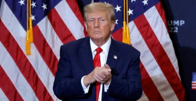 Donald Trump in a blue suit with a red tie claps his hands warily in front of American flags