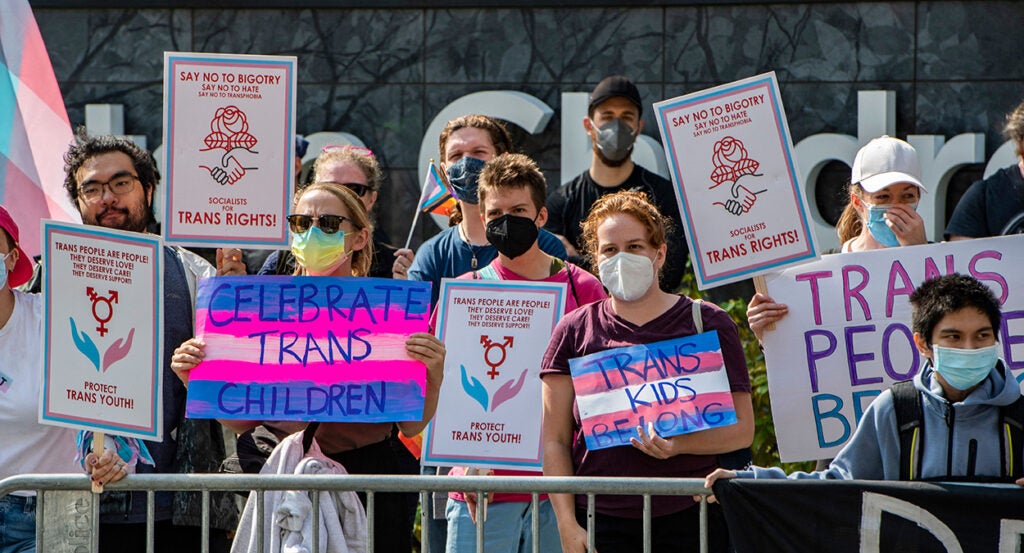 Protesters wearing masks hold signs reading "Celebrate trans children" and "trans kids belong"