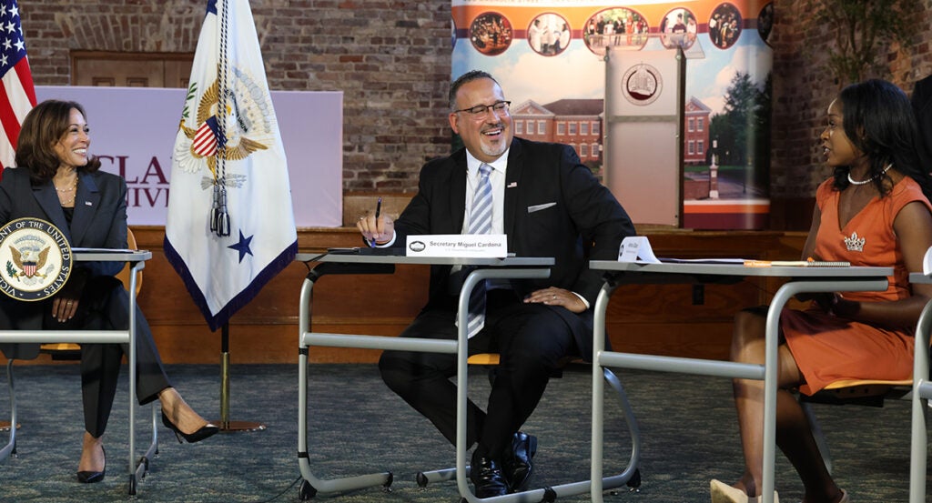 Kamala Harris in a suit, Miguel Cardona in a suit, sit behind desks