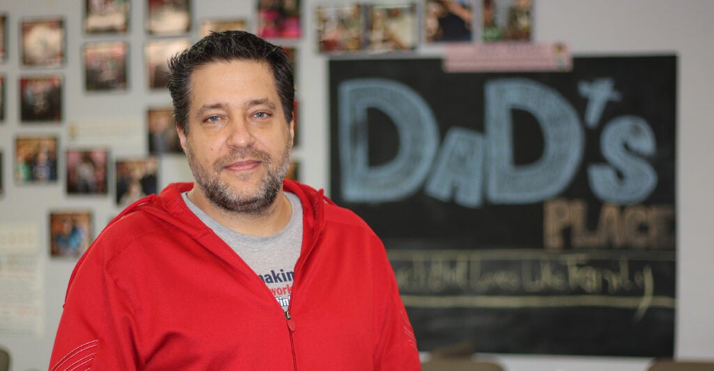 Pastor Chris Avell stands in front of a church bulletin board wearing a red sweatshirt.