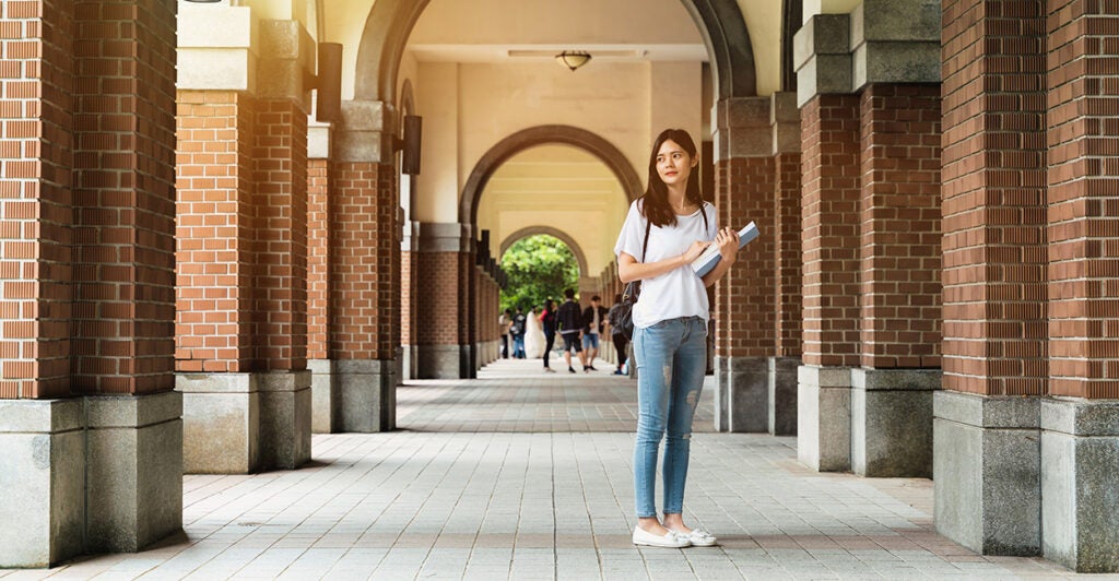 A girl stands in a passageway on a college campus with people in the distance behind her.