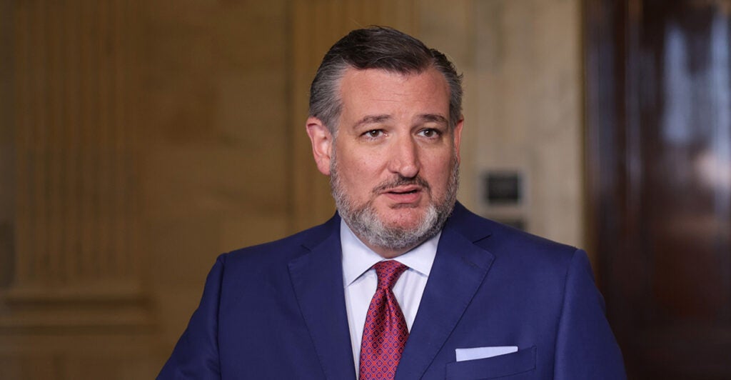 Sen Ted Cruz standing in a blue suit and red tie while speaking to members of the press.