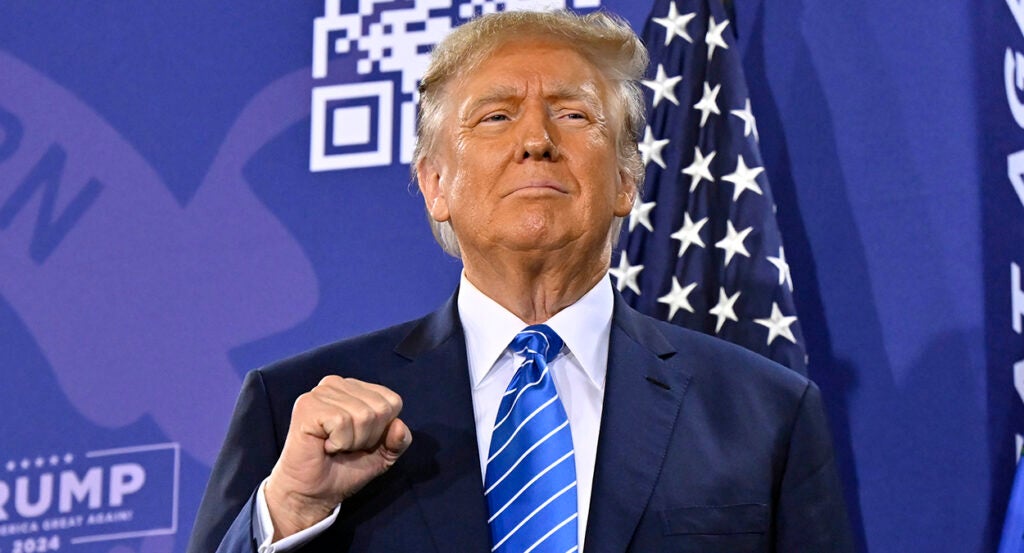 Donald Trump raises a fist with a grim smile on his face, wearing a blue suit in front of an American flag