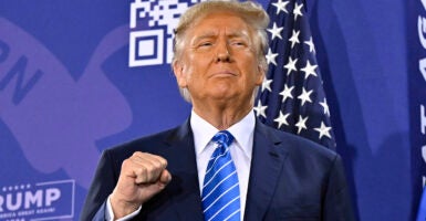 Donald Trump raises a fist with a grim smile on his face, wearing a blue suit in front of an American flag