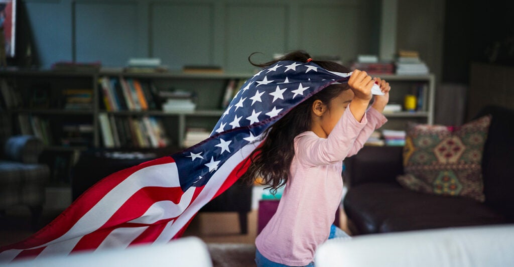 A little girl runs through her home with an American flag.