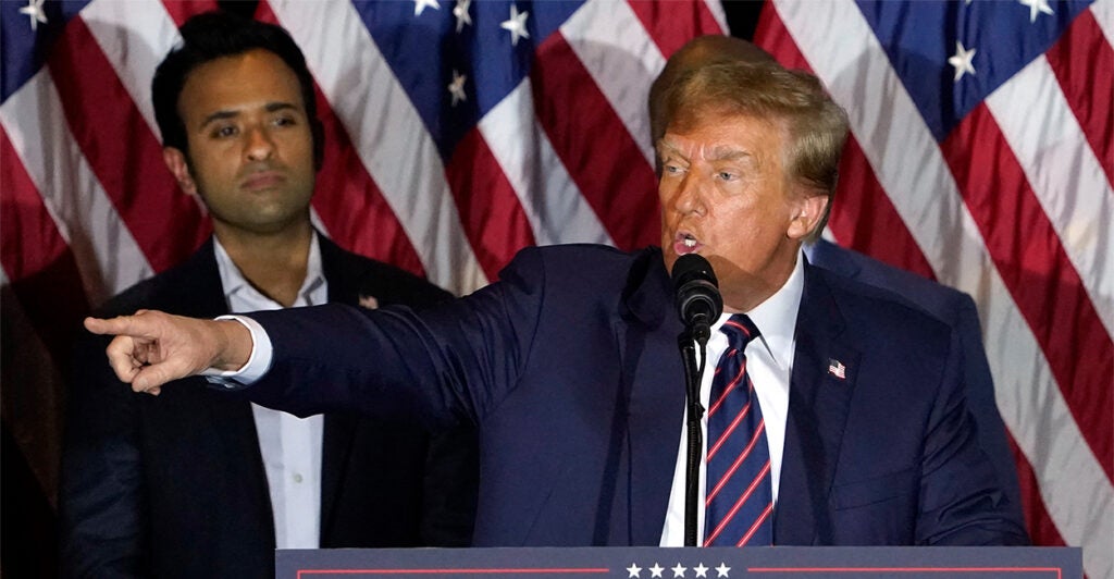 Trump stands at a podium pointing with a row of American flags behind him. Vivek Ramaswamy stands to Trump's left looking on.