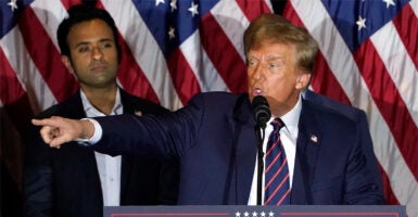 Trump stands at a podium pointing with a row of American flags behind him. Vivek Ramaswamy stands to Trump's left looking on.