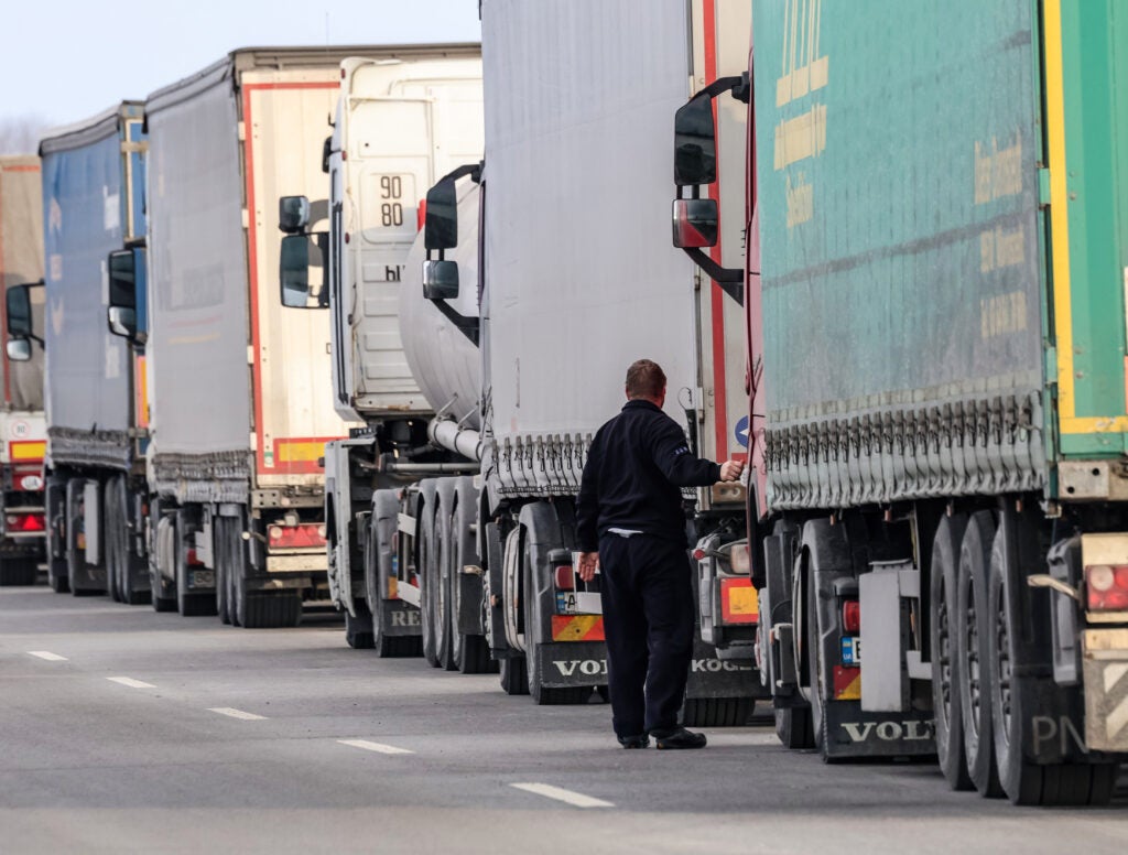 Trucker in black outfit checks truck as trucks queue across border.