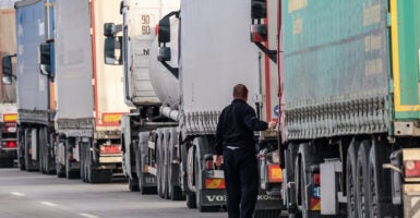 Trucker in black outfit checks truck as trucks queue across border.