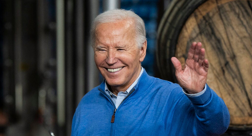 President Joe Biden waves in a blue sweatshirt in front of a beer barrel