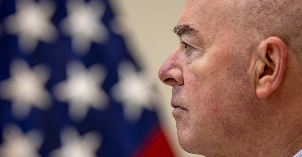 Side profile view shows DHS Secretary Alejandro Mayorkas looking ahead with an American flag behind him.