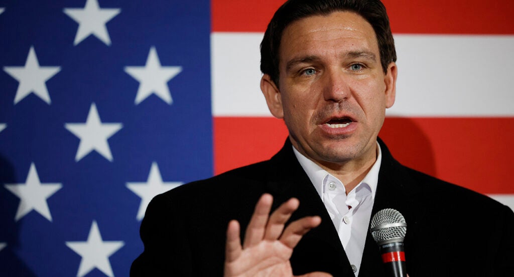 Ron DeSantis in a black suit stands in front of an American flag