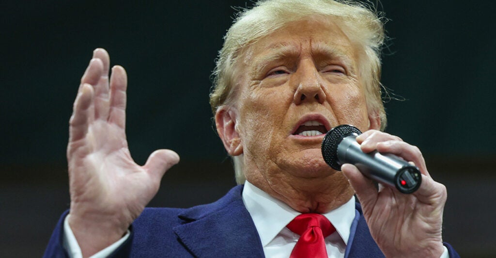 Former President Donald Trump speaks into a microphone in a blue suit and red tie.