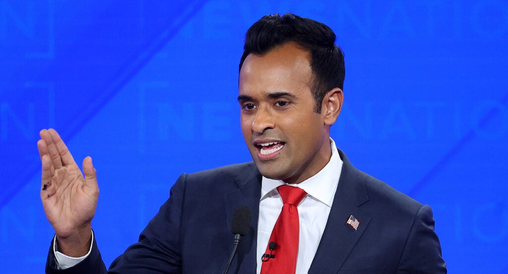 Vivek Ramaswamy gestures in a black suit with a red tie and an American flag pin