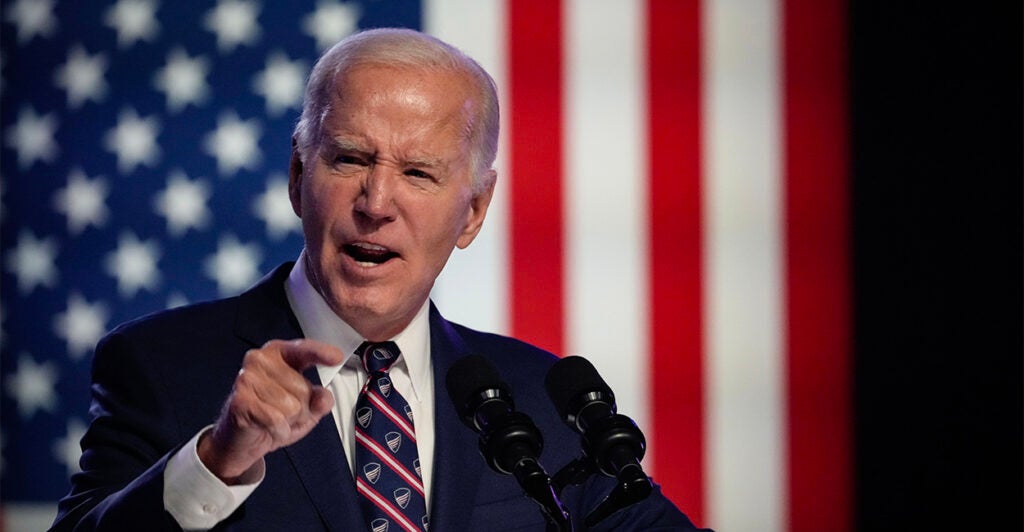 Joe Biden speaks at a podium in a suit with a large flag hanging behind him