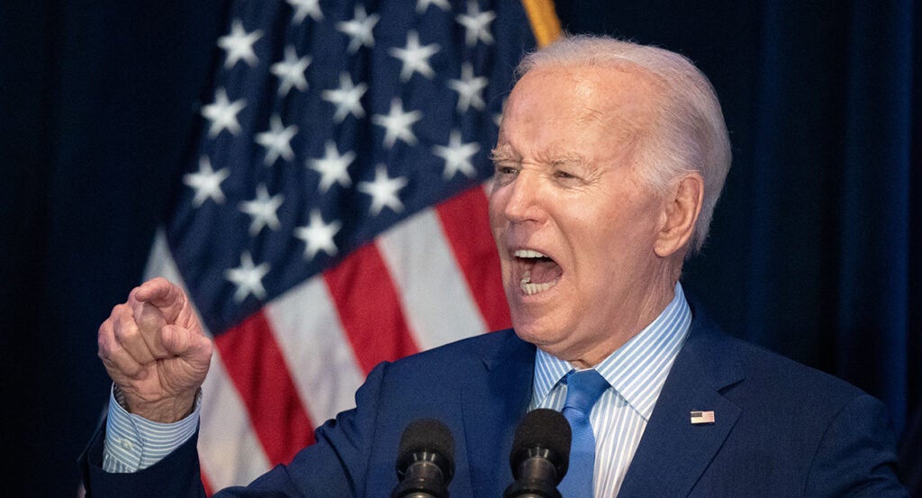 Joe Biden in a blue suit gestures angrily in front of an American flag while wearing an American flag pin.