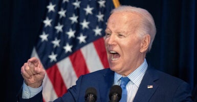 Joe Biden in a blue suit gestures angrily in front of an American flag while wearing an American flag pin.